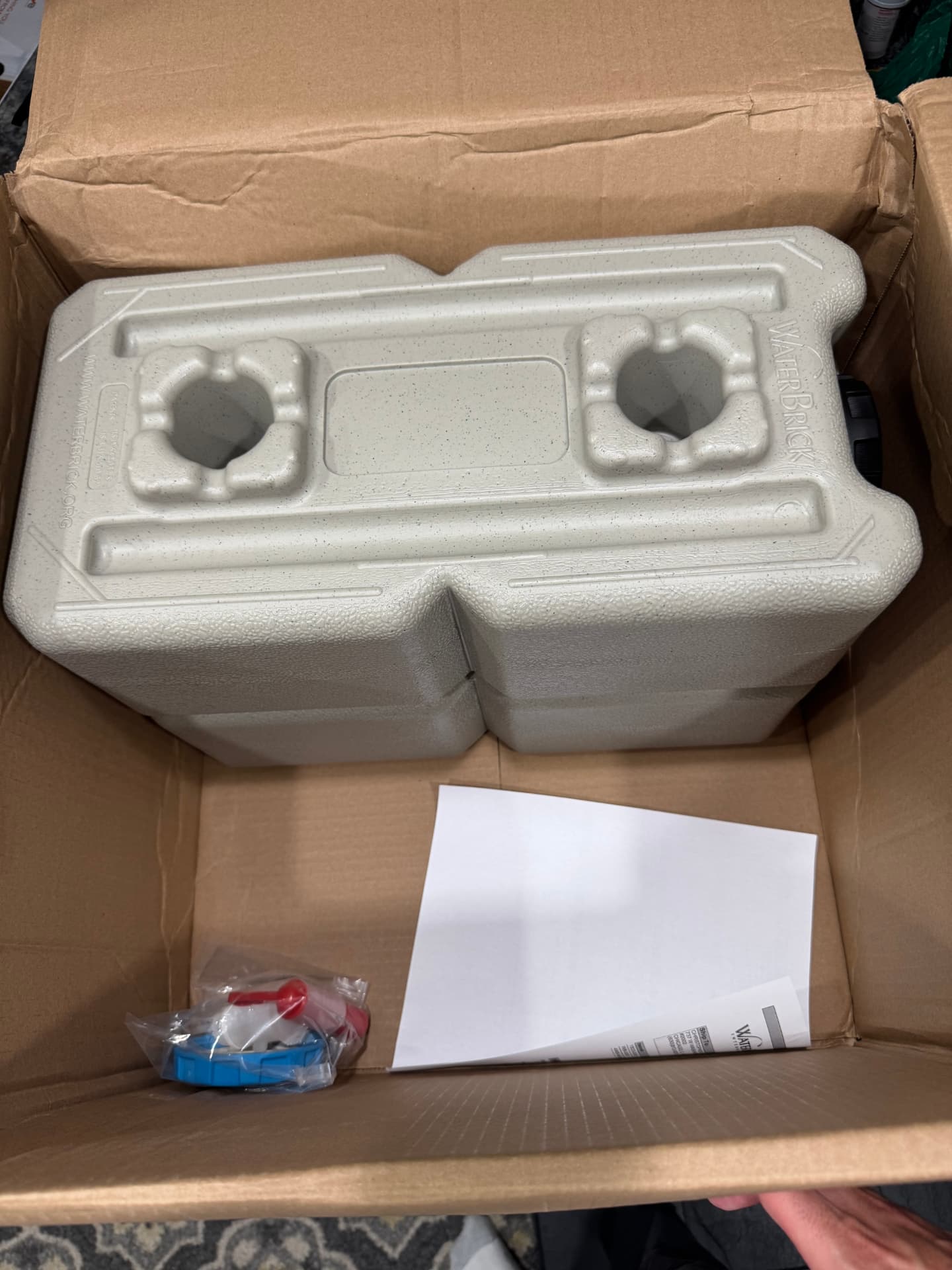 An earth-tone WaterBrick container sitting in an oversized shipping box with a bagged spigot kit and paperwork