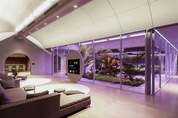 Interior of a modern underground shelter with curved white ceilings, an air quality display and a glass-walled hydroponic garden lit in violet