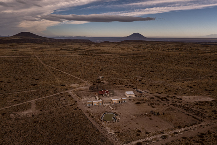 Aerial view of a remote high-desert compound with low buildings, dirt tracks and a reservoir, surrounded by empty scrubland and distant volcanic peaks